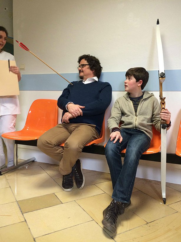 Man and son at doctor's office, man has arrow in his chest and kid is holding a bow.