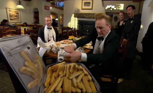 Conan O'Brian filling up a suitcase with breadsticks.