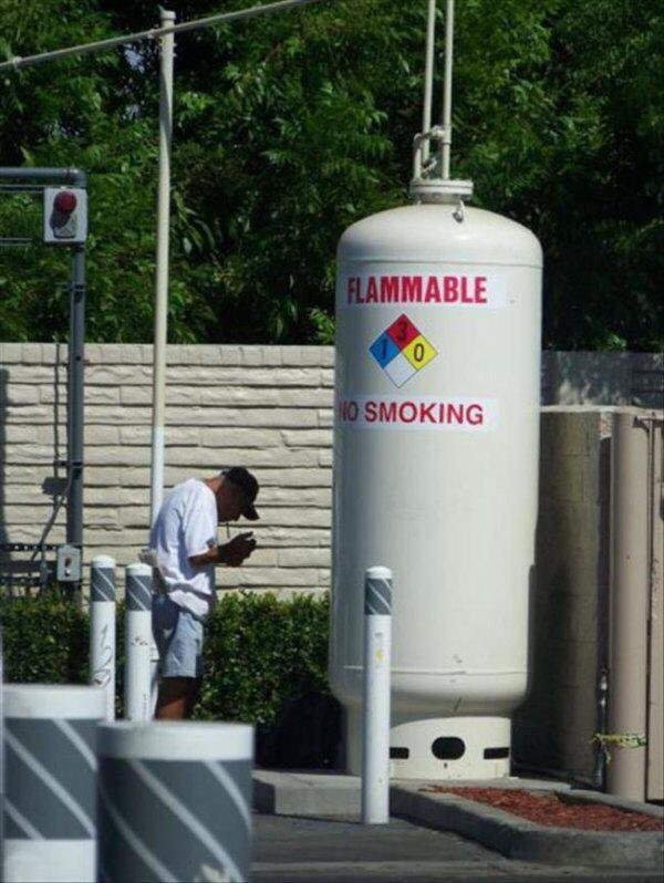 funny safety workplace fails - man lighting cigarette near giant gas tank that says flammable no smoking