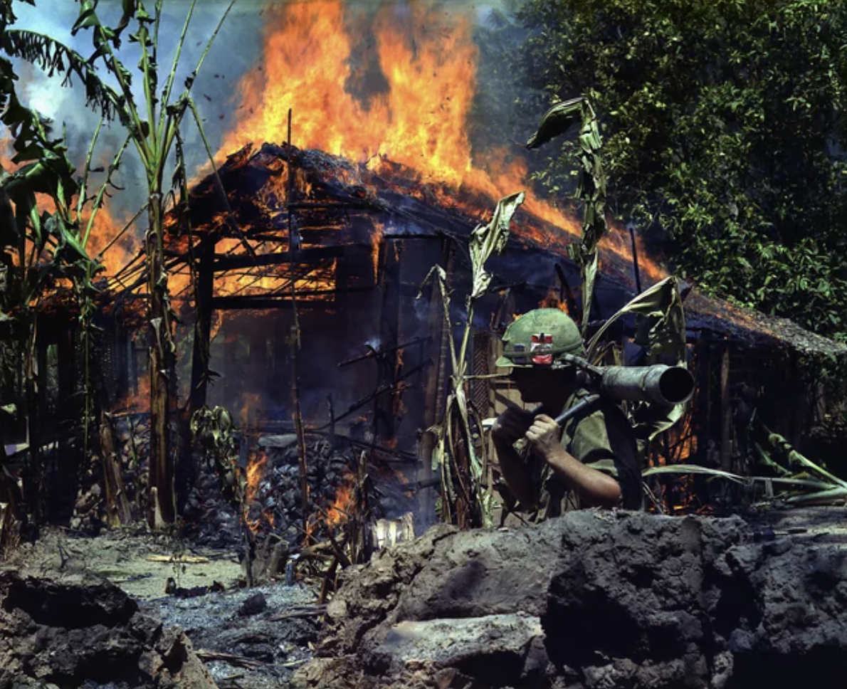 A Viet Cong base camp being burned during the Vietnam War. An American private first class stands by. April 5th, 1968.
