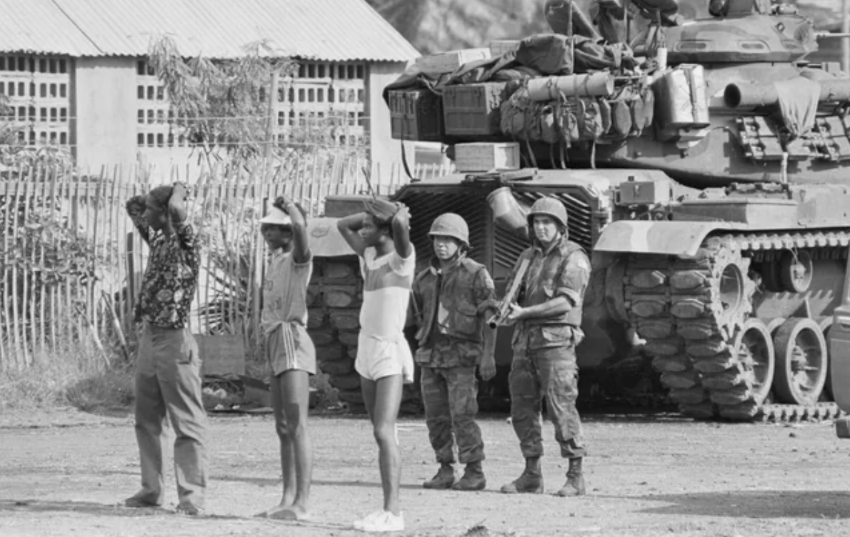 Two U.S. soldiers hold three suspected members of the People's Revolutionary Army at gunpoint in St. George's, Grenada during the Grenada Invasion in 1983.