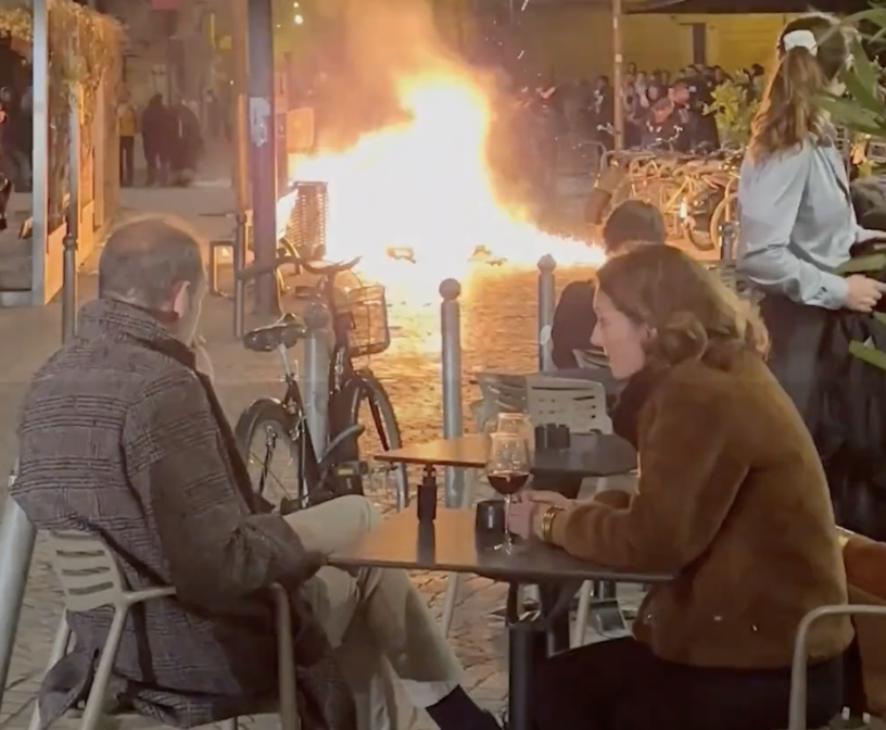 French couple sips wine while fires burn from protests in the background. 