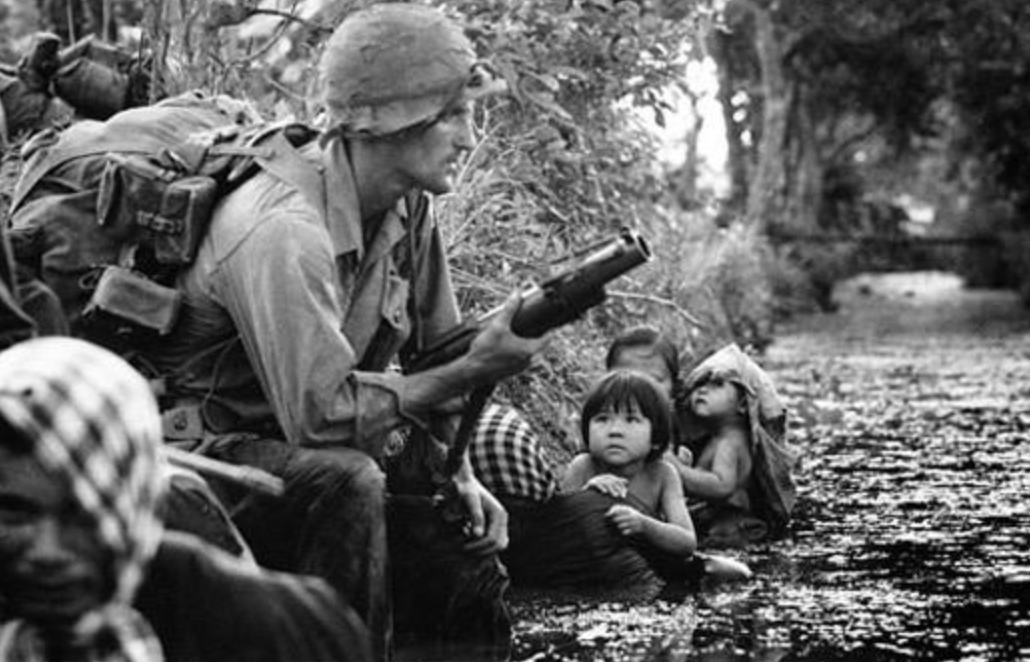 Two Vietnamese children, huddling against a canal bank for protection from Viet Cong sniper fire, gaze at an American paratrooper's M79 grenade launcher on New Year's Day, 1966, near Bao Trai.