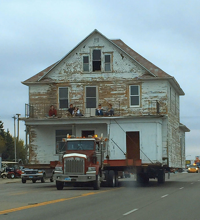 A house being moved down the interstate with people sitting on the 2nd storey porch.