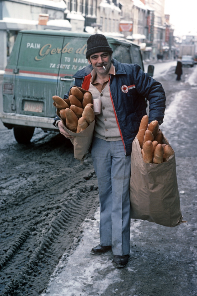 A bread delivery man with bags filled with baguettes on a snowy street in Quebec in 1977.