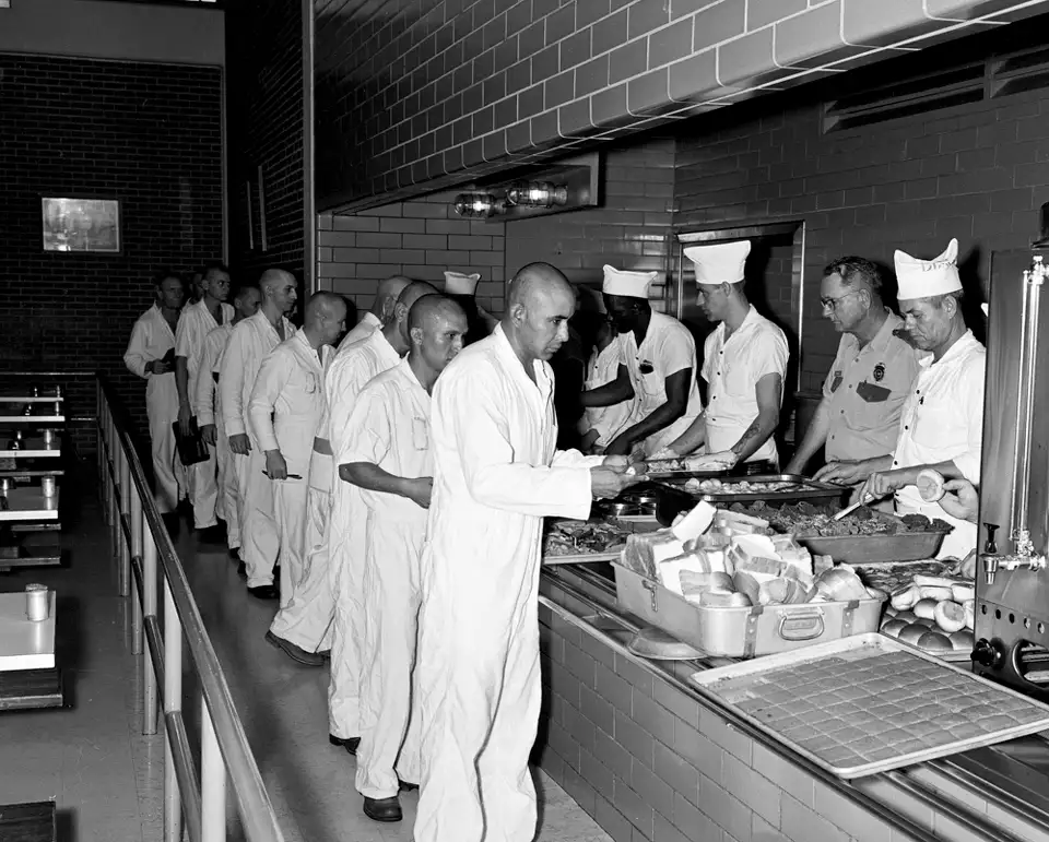 Prisoners dressed in white uniforms wait their turn in the hall, trays in hand, overseen by guards and kitchen staff.