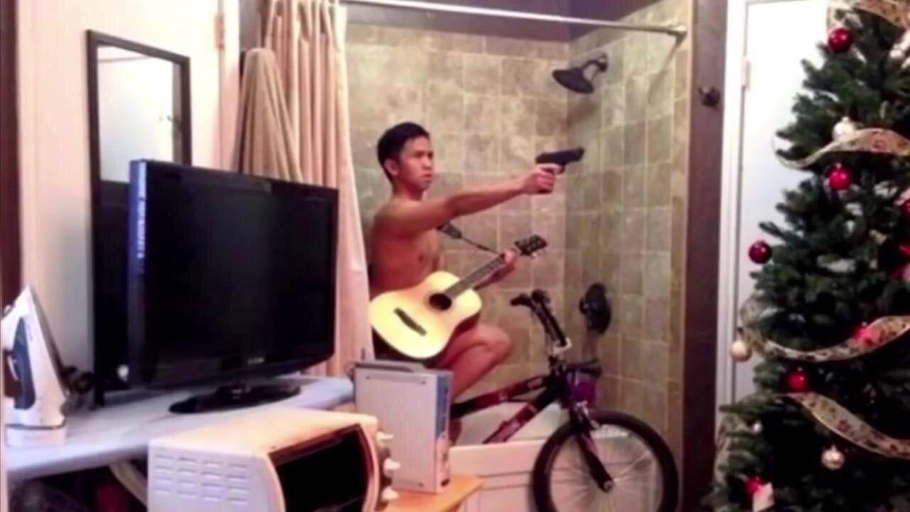 man riding a bike in his bathtub while playing guitar and pointing a gun with TV and christmas tree also present in the room