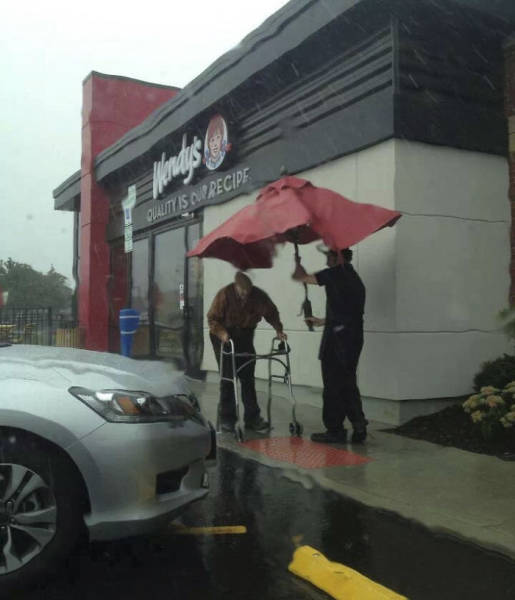 A Wendy's employee removing an umbrella from a table outside to walk an elderly gentleman to his car in the rain.