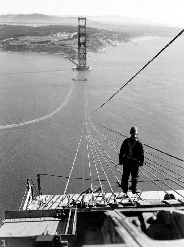 A man standing on the first cables during the construction of the Golden Gate Bridge, with the Presidio and San Francisco in the background (1935)