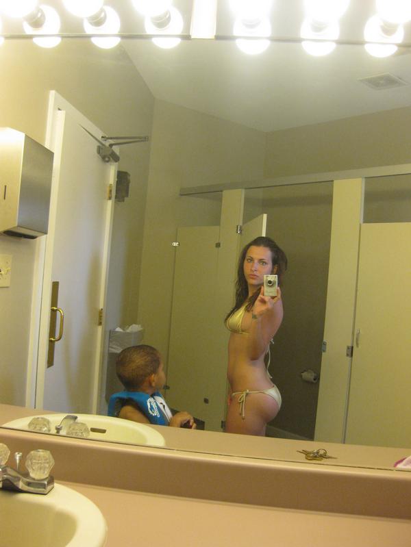 Girl takes picture of herself in a bikini in public restroom of a inner city pool while her kid watches.