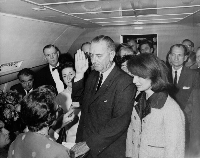 Jacqueline Kennedy wears her pink Chanel suit, still stained with the blood of her husband, as Lyndon Johnson takes the oath of office in Air Force One. According to Lady Bird Johnson, who was also present: "Her hair was falling in her face but she was very composed ... I looked at her. Mrs. Kennedy's dress was stained with blood. One leg was almost entirely covered with it and her right glove was caked, it was caked with blood  her husband's blood. Somehow that was one of the most poignant sights  that immaculate woman, exquisitely dressed, and caked in blood."