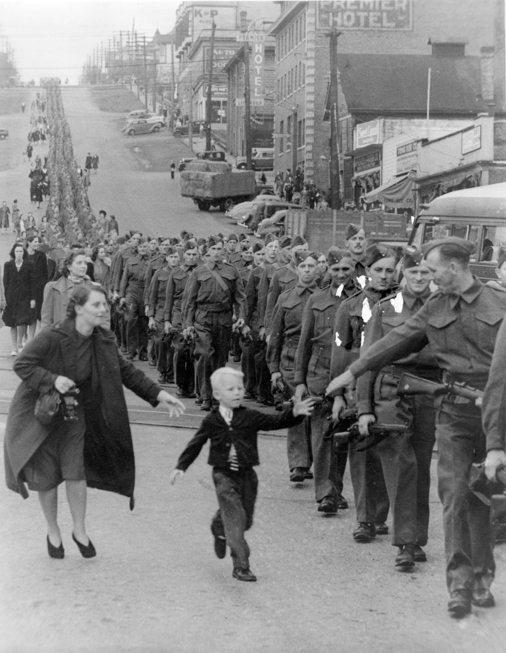 “Wait For Me Daddy,” by Claude P. Dettloff, October 1, 1940: A line of soldiers march in British Columbia on their way to a waiting train as five-year-old Whitey Bernard tugs away from his mother’s hand to reach out for his father.