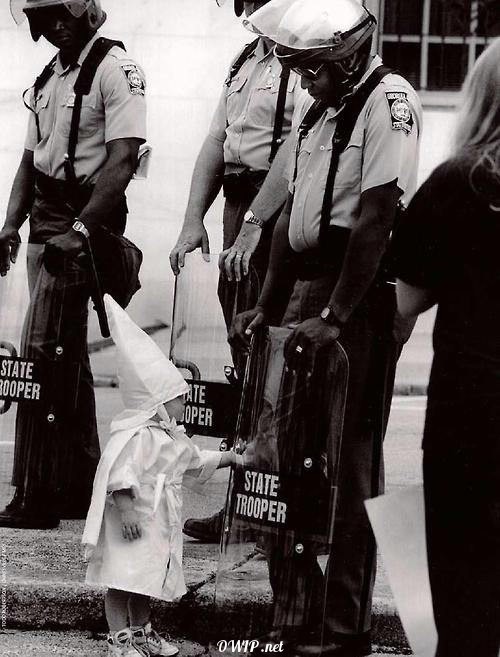 Child dressed in Ku Klux Klan outfit, playing with his reflection on the shield of an African-American police officer during a KKK demonstration.