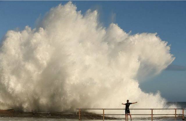 Man at the coast taking it all in with giant wave about to hit him with a dose of reality