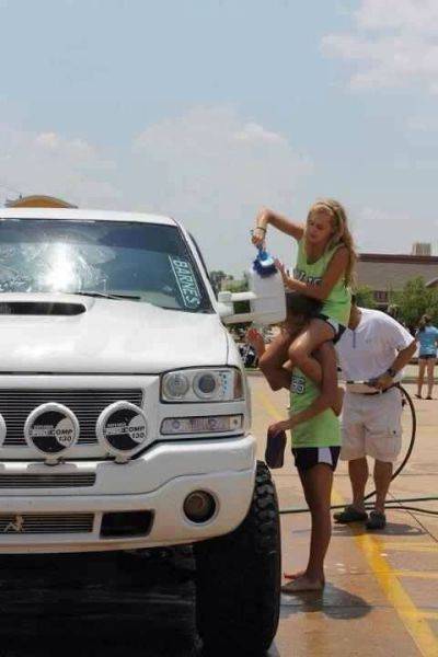 Picture of a girl on a man's shoulders to clean the top of the mirror on a pickup truck.