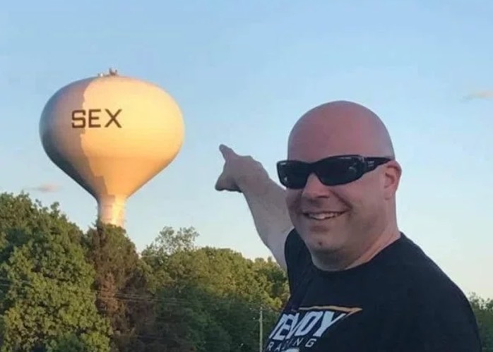 Man pointing at a water tower with a word on it that starts with S