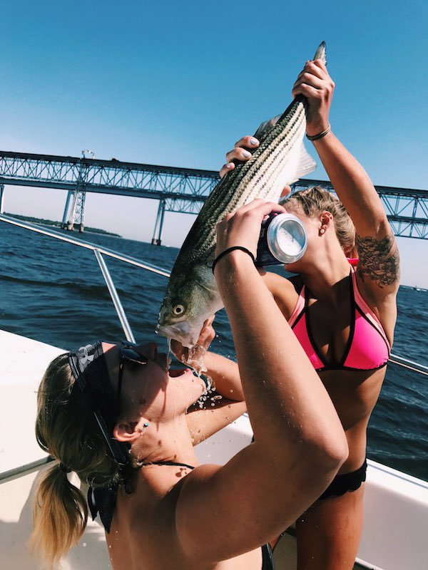 Two girls in bikinis on a boat drinking beer out of a fish.