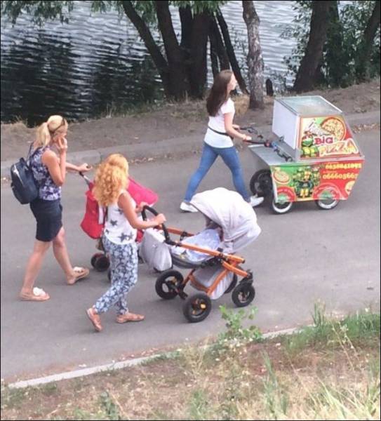 Women pushing their carriages in the park, one of them is not a baby but a pizza wagon.