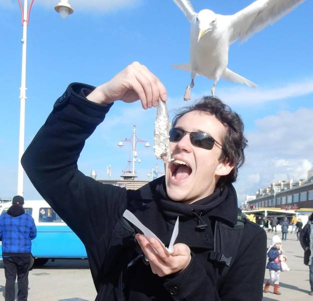 Man about to eat some fish by the seaside and a seagull is about to snatch it.