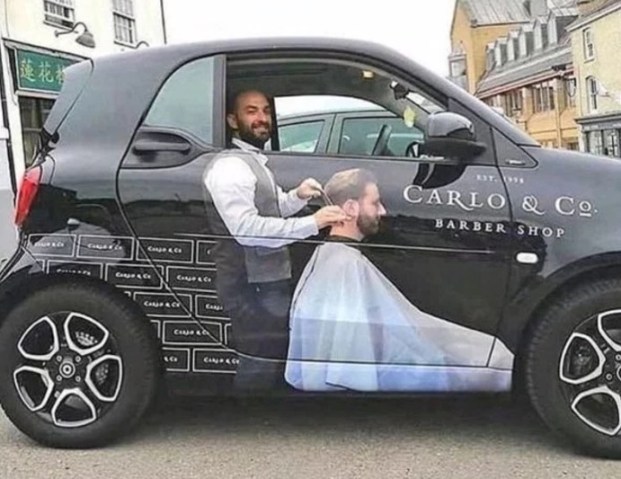 Barber with a car that makes it look like he is working because of the image on the side of the car.