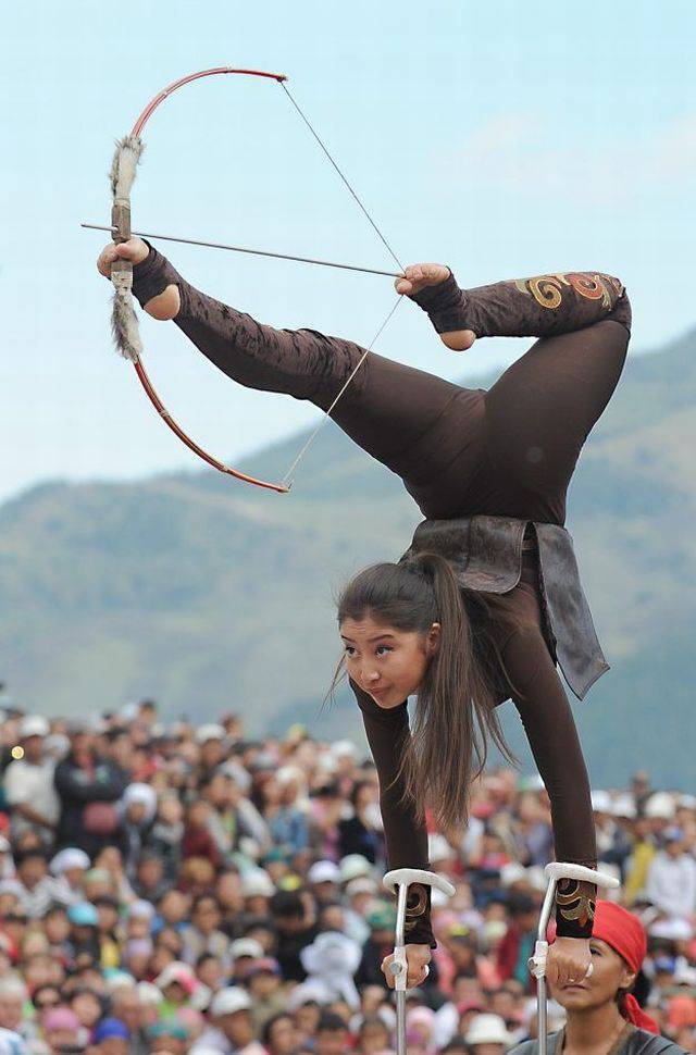 Girl standing on her hands and shooting bow and arrow with her feet.
