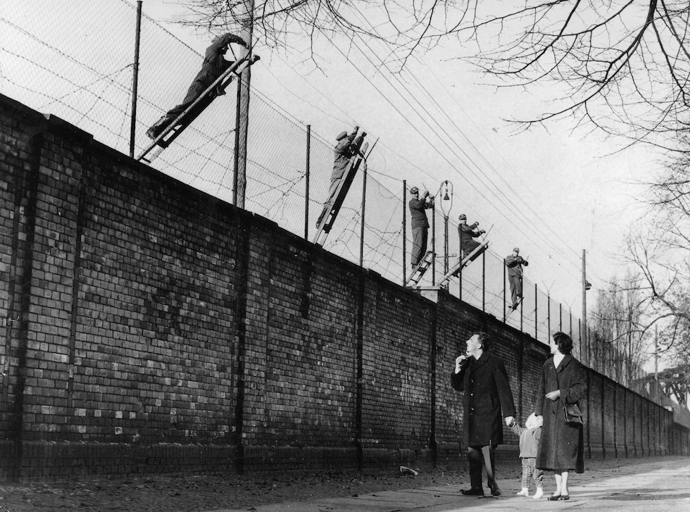 East Berlin guards adding fresh barbed wire to the top of part of the Berlin Wall in 1972 as a curious West German family watches.