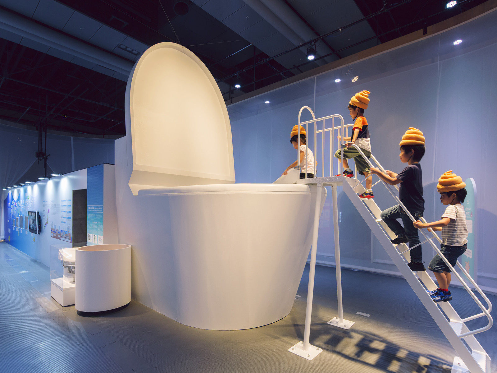 Japanese children wearing feces-shaped hats line up to slide down into a giant toilet, simulating the magical journey of human waste matter through a virtual sewer world, at a toilet exhibition in Tokyo