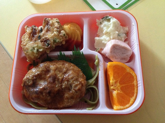 Country: Japan. Contents: Tofu hamburger with pasta, fishcake, fried potato with ketchup, some kind of potato salad, wiener, and mikan tangerine.