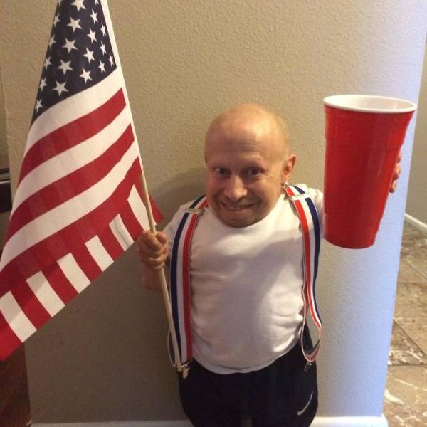 Verne Troyer (mini-me) holding up a large beer cup, an american flag, and red-white-blue suspenders.