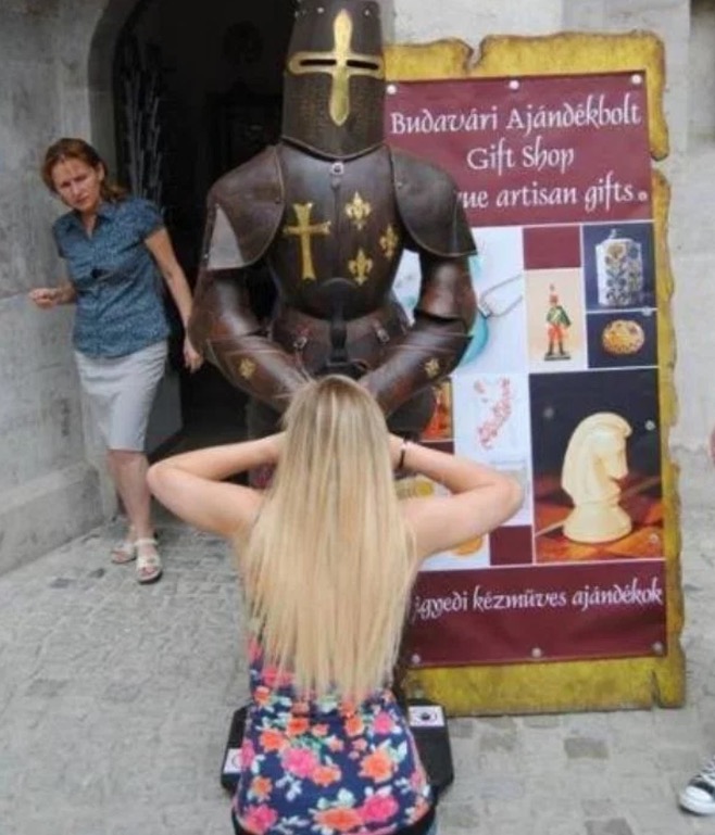 Woman crouching in front of Knight In Armor to look like she is giving it a BJ, when she is just posing for the camera.