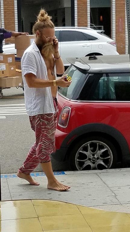Bearded hipster with a man bun walking the street barefoot while on the phone and fidgeting a Rubik's cube.