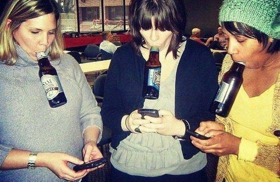 Girl holding beer bottles in their mouth as they thumb their phones.