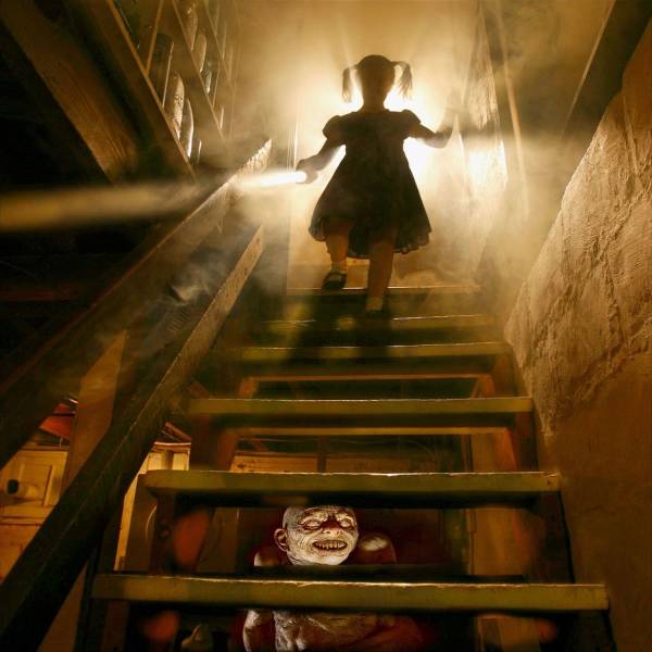 creepy cursed picture of a girl coming down the stairs with light behind her and a poster of Pennywise from IT or some other clown on a poster beyond the stairs