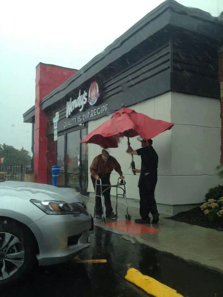 An employee removes an outdoor seating umbrella to shelter an elderly man as he walks to his car.