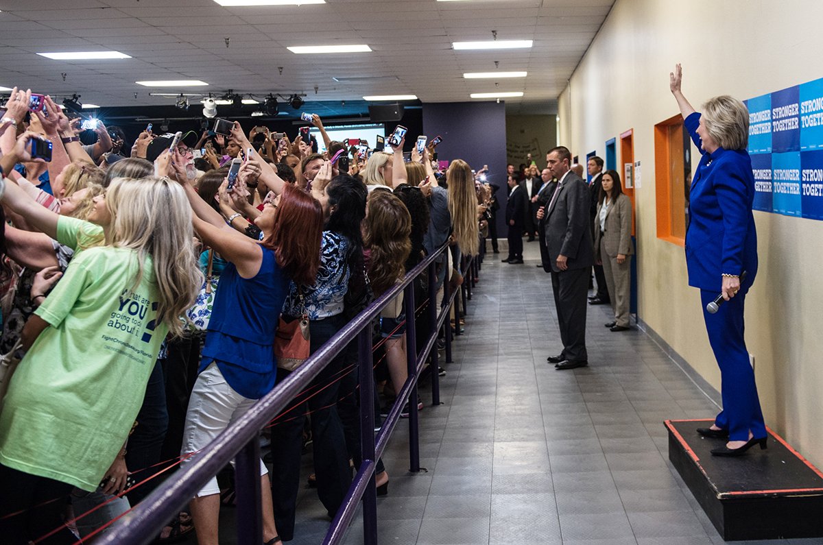 As Hillary Clinton stands on a podium and waves, everyone has turned their backs and raised their phones in order to get a little of themselves...