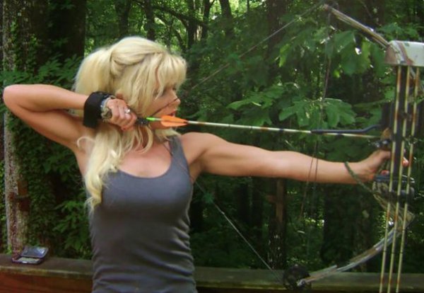 Muscular woman in a tank top shooting a bow and arrow.