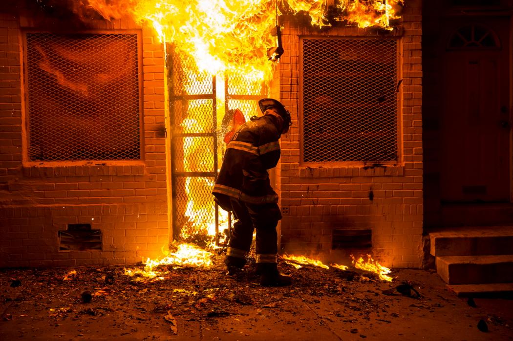 A firefighter opening a gate with a saw while fending off fire at a convenience store during the Baltimore riots.
