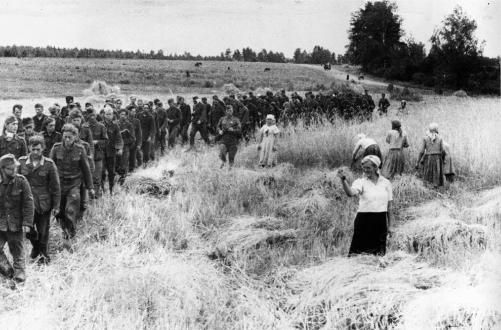 German prisoners of war marching east under the supervision of Soviet soldiers, 1944. Take note of the Soviet woman showing a 'dulya' with her hand to the Germans as they pass by.
