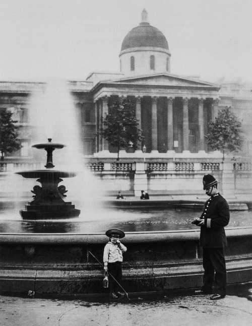 A policeman questions a young boy who probably intended to fish in the fountain on Trafalgar Square, London, 1892