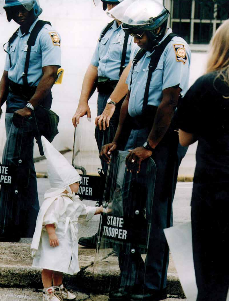 A young boy in KKK robes sees his reflection in a riot shield held by a Georgia state trooper. Gainesville, Ga., in 1992