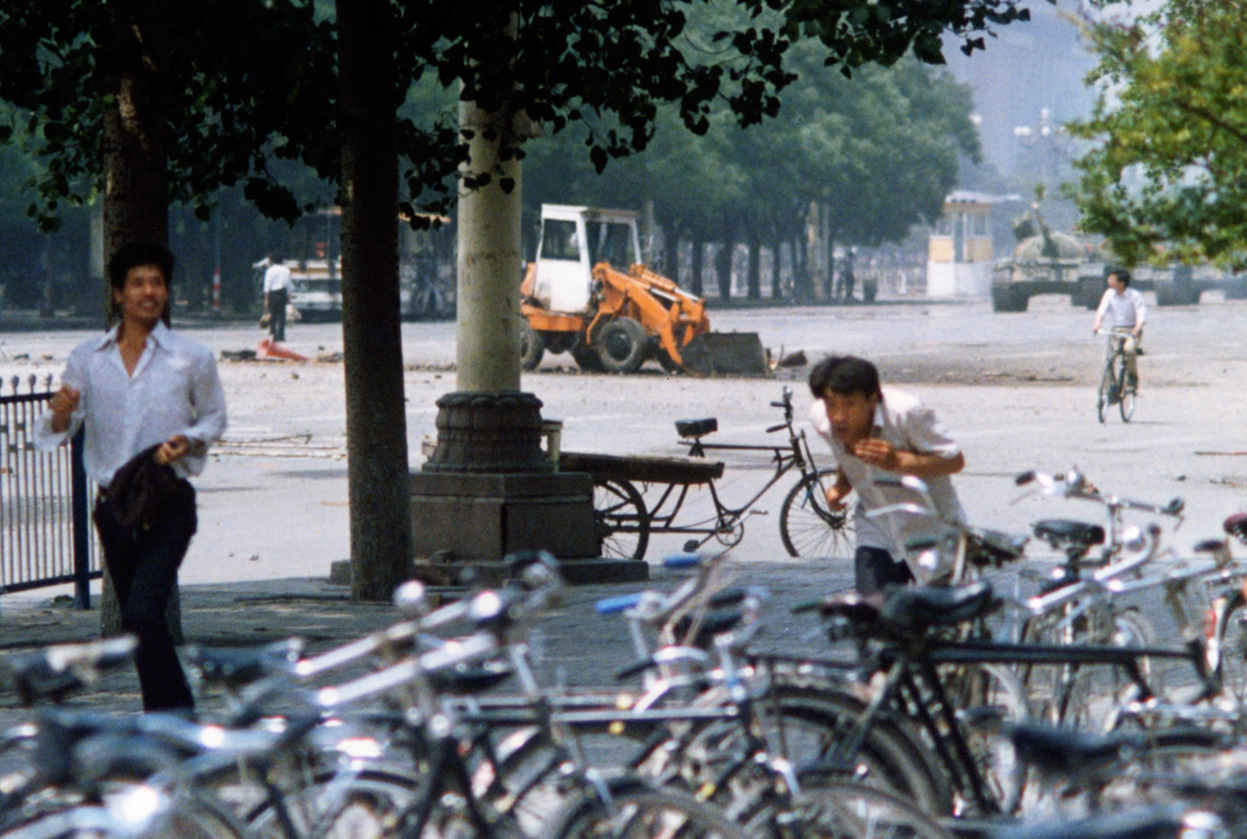 A different angle taken of "Tank Man," the man who stood against a line of tanks in Tiananmen Square. He is standing in the street between the tree trunk and the fleeing man. You can see the tanks approaching from the right.