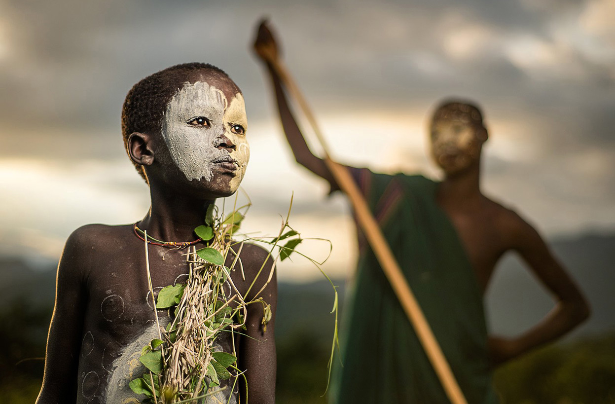 A BOY OF THE NOMADIC SURI TRIBE OF ETHIOPIA, IN TRADITIONAL FACEBODY PAINT AND ATTIRE
