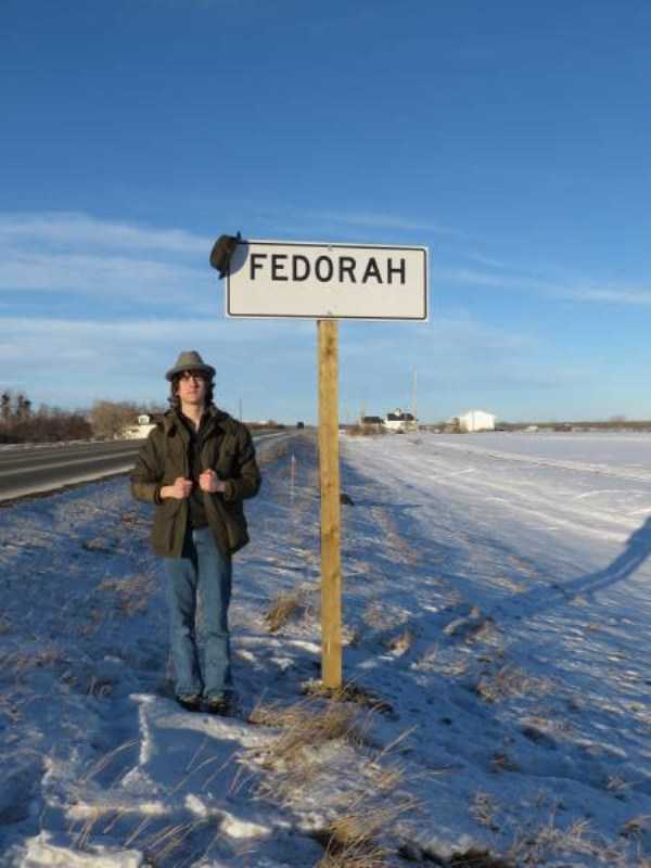 Cringe of a man wearing a fedora and also a fedora hang up on the sign he is next to which is to the town of Fedorah.