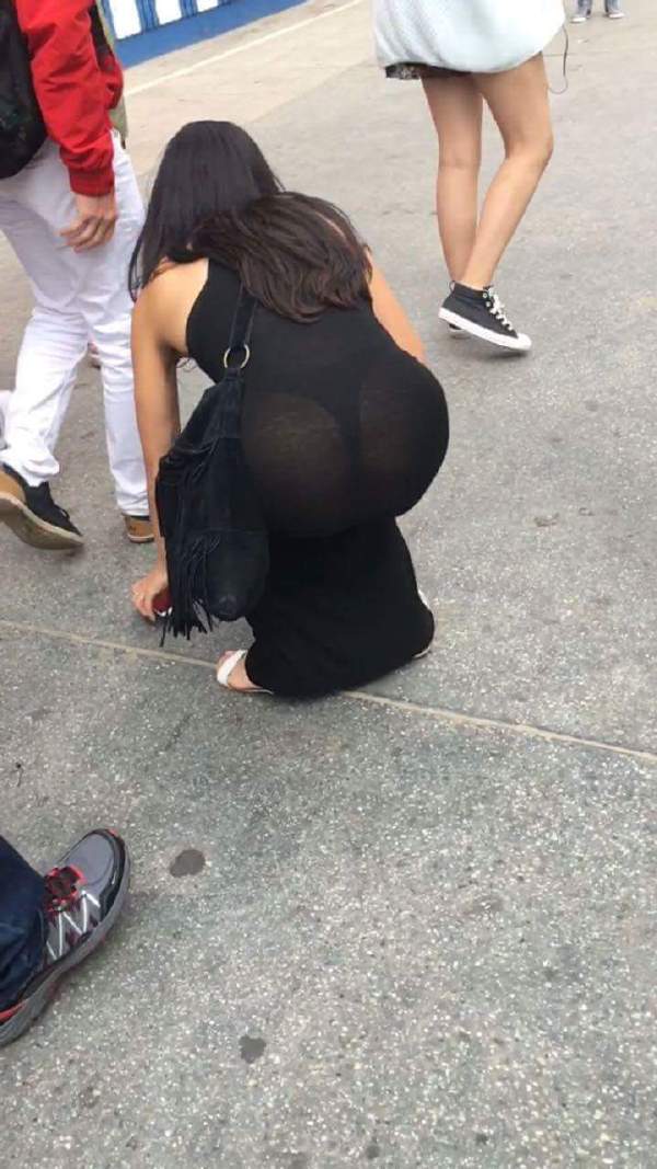 Pic of woman in black dress that is partially see thru bending down to pick up something on the sidewalk.