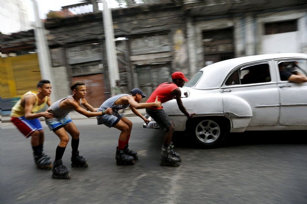 Teenagers on roller skates hold on to each other as they are pulled by a vintage car to move along a street in Havana