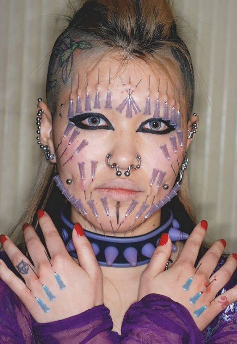 Picture of a girl with piercings and many medical needles stuck into her face and hands.