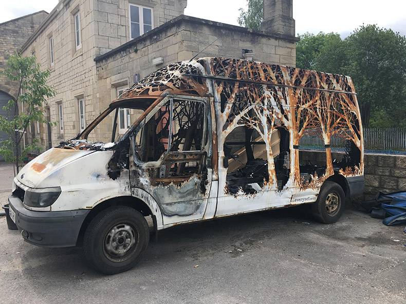 Burnt out van with trees cut into the remaining shell of the vehicle.