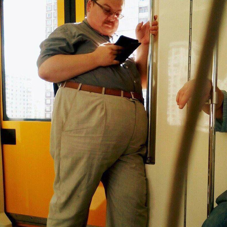 Really fat man with pants pulled up high riding the subway and reading his phone.