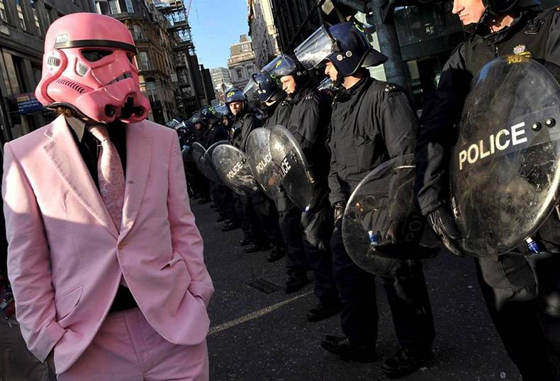 Man in a pink suit wearing a pink Darth Vader outfit with hands in pocket and staring down a bunch of police officers in riot gear.