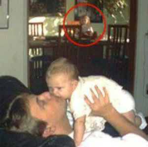 Man kissing his kid in an old photo with a mysterious character lurking in the background.